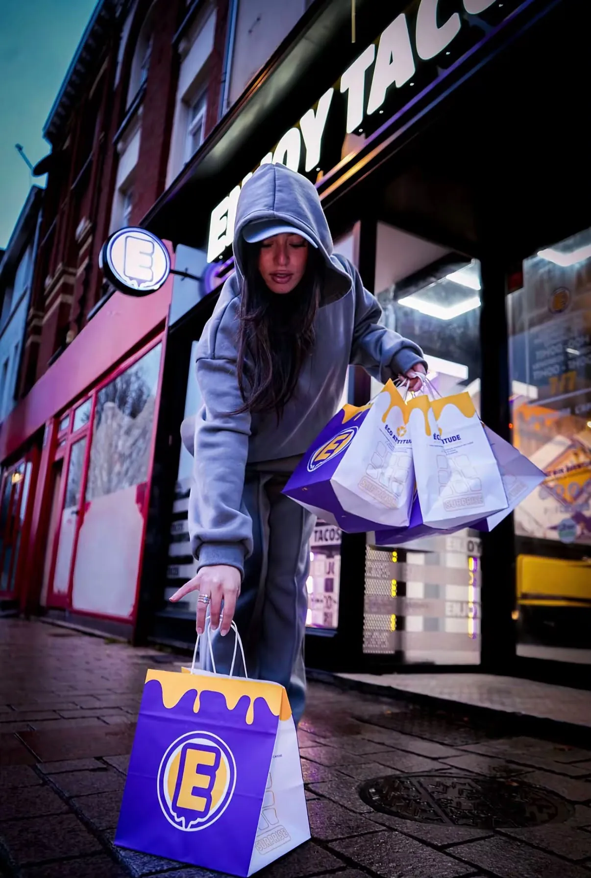 Une jeune femme en survêtement dépose des sacs Enjoy Tacos devant un restaurant, en pleine rue