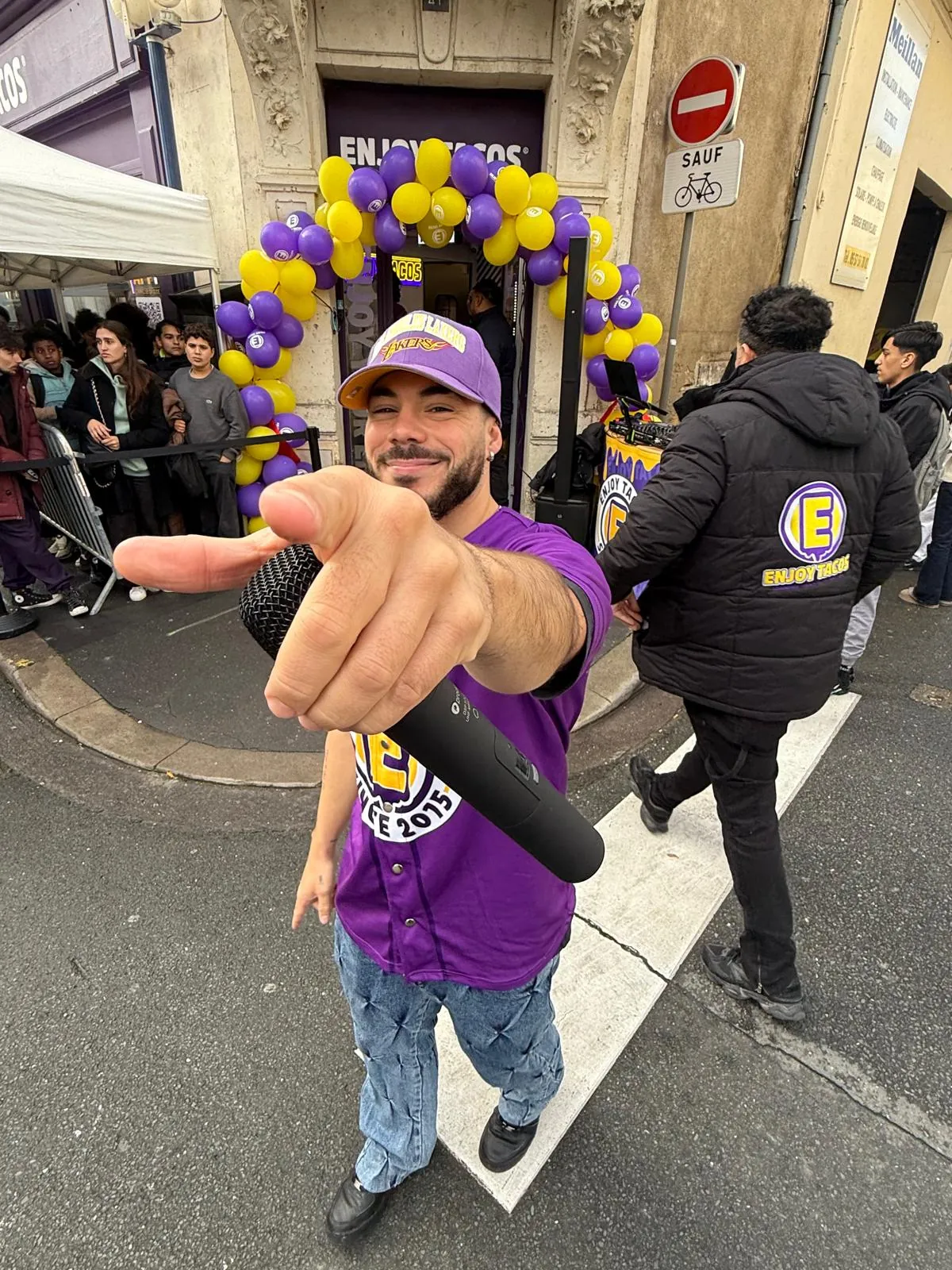 Inauguration d’un restaurant Enjoy Tacos : un animateur en maillot violet devant l’arche de ballons violets et jaunes, public massé derrière les barrières