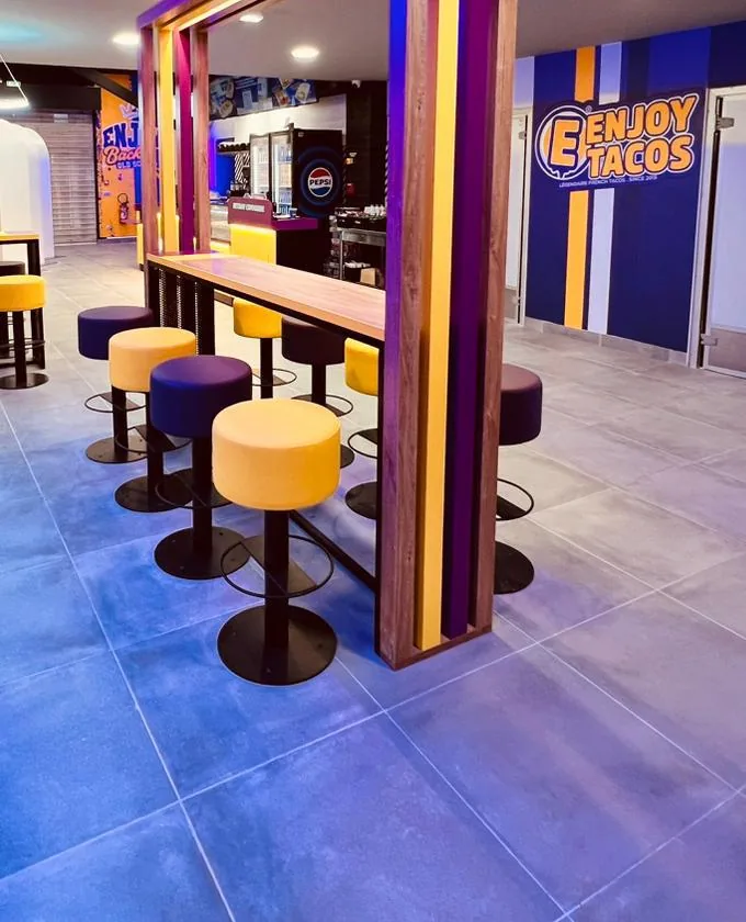Détail de la salle : tabourets violets et jaunes autour d’un cadre en bois lamellé